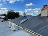 Roof with solar panel, air conditioning unit, and chimneys under a blue sky with clouds.