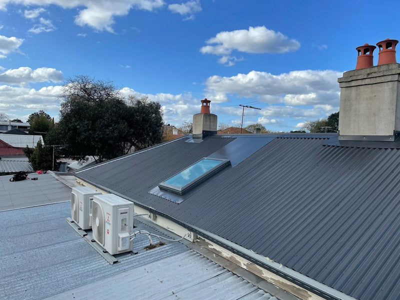 Roof with solar panel, air conditioning unit, and chimneys under a blue sky with clouds.