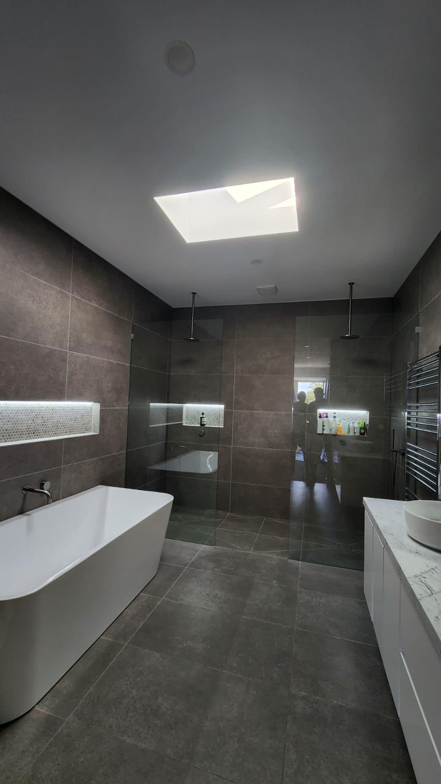 Modern bathroom with dark tiled walls, a white freestanding bathtub, and a skylight. - Bill Roofing 