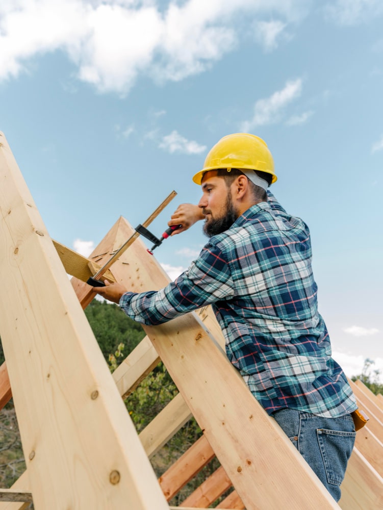 Man in a hard hat and plaid shirt working on a wooden structure against a blue sky.